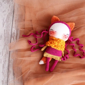May include: A handmade crochet doll with a fox-like design. The doll features a white face, pink cheeks, and closed eyes. It wears a pink dress, an orange scarf, and has orange fox ears. Long, curly pink yarn strands extend from the head.
