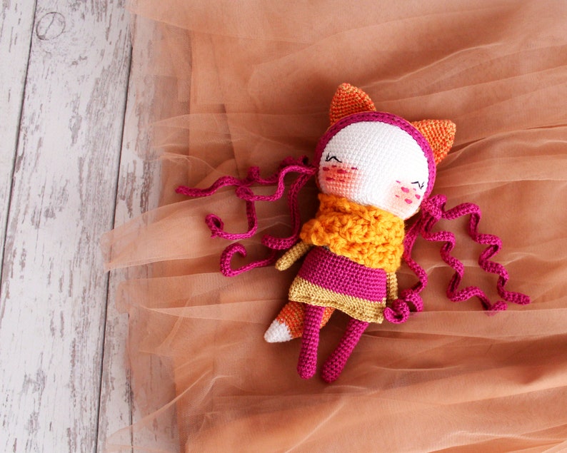May include: A handmade crochet doll with a fox-like design. The doll features a white face, pink cheeks, and closed eyes. It wears a pink dress, an orange scarf, and has orange fox ears. Long, curly pink yarn strands extend from the head.