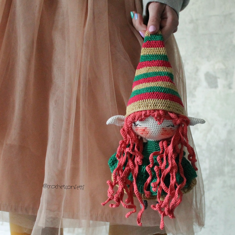 Peut inclure: Une poup&eacute;e elfe en crochet avec une robe verte, des cheveux roux et un chapeau ray&eacute; rouge et vert. La poup&eacute;e est tenue par une personne portant une jupe en tulle rose.
