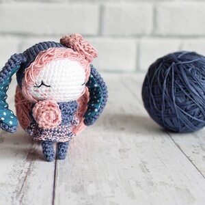 K&ouml;nnte beinhalten: Ein geh&auml;keltes Amigurumi-Spielzeug in Form eines Hasen mit blau-wei&szlig;en Polka-Dot-Ohren, einer rosa Blume auf dem Kopf und einer rosa Blume in der Hand. Der Hase tr&auml;gt ein blau-rosa Kleid und hat geschlossene Augen.