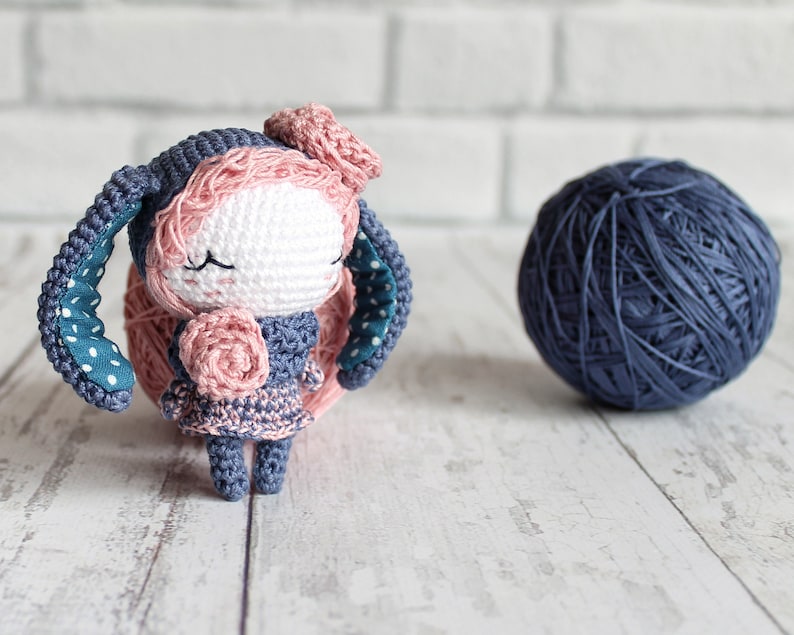 K&ouml;nnte beinhalten: Ein geh&auml;keltes Amigurumi-Spielzeug in Form eines Hasen mit blau-wei&szlig;en Polka-Dot-Ohren, einer rosa Blume auf dem Kopf und einer rosa Blume in der Hand. Der Hase tr&auml;gt ein blau-rosa Kleid und hat geschlossene Augen.
