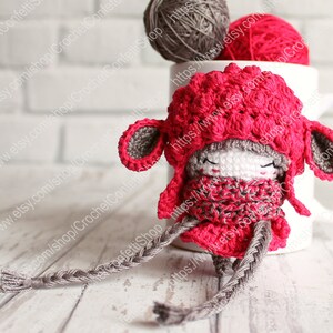 May include: A small crocheted doll wearing a red hat and scarf. The doll has a braided gray yarn tail and is sitting on a white wooden surface.