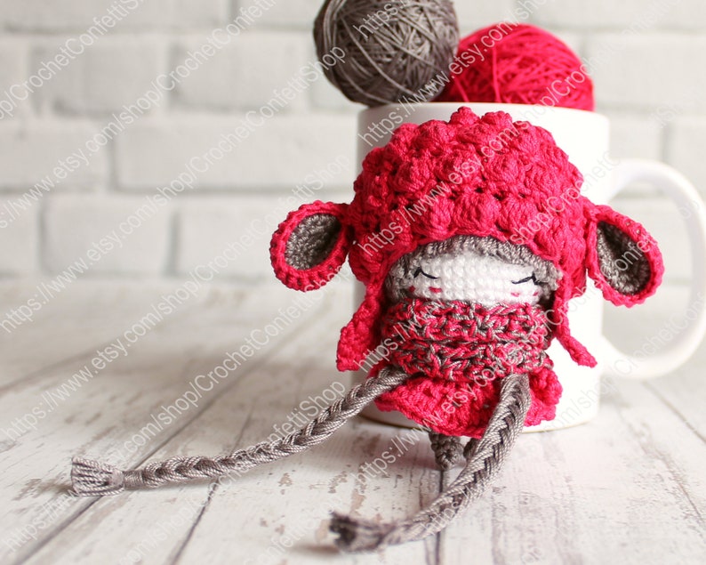 May include: A small crocheted doll wearing a red hat and scarf. The doll has a braided gray yarn tail and is sitting on a white wooden surface.