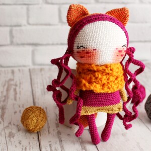 May include: A handmade crochet doll with a white face, rosy cheeks, and closed eyes. The doll has orange cat ears, curly magenta hair, and a matching scarf. The doll is wearing a magenta dress with a gold trim and magenta legs.