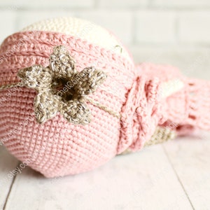Op de afbeelding: Een roze en cr&egrave;me gebreide baby muts met een bruin gebreid bloemenaccent. De muts is gemaakt van zacht garen en is perfect voor een pasgeboren baby.