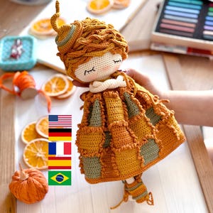 Op de afbeelding: Een gehaakte pop met bruin haar, gesloten ogen en een patchworkjurk in tinten bruin en groen. De pop draagt een witte gehaakte kraag en een bruine gehaakte hoed.