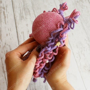 Puede incluir: Una mu&ntilde;eca de crochet rosa con pelo largo de hilo morado y rosa. La mu&ntilde;eca est&aacute; siendo sostenida por las manos de una persona.