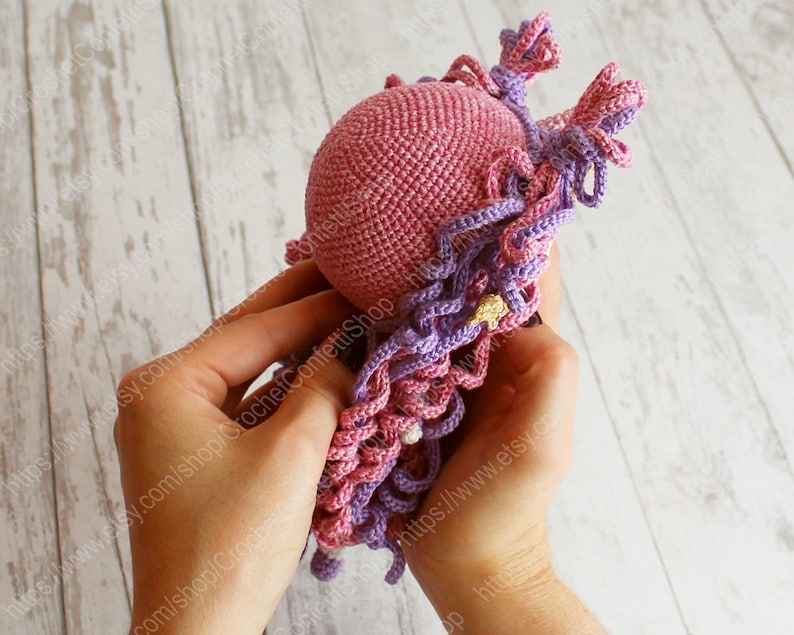 Puede incluir: Una mu&ntilde;eca de crochet rosa con pelo largo de hilo morado y rosa. La mu&ntilde;eca est&aacute; siendo sostenida por las manos de una persona.