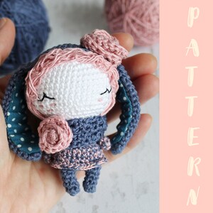 K&ouml;nnte beinhalten: Eine geh&auml;kelte Puppe aus rosa und blauem Garn. Die Puppe hat ein wei&szlig;es Gesicht mit geschlossenen Augen und eine rosa Blume auf der Brust. Die Puppe h&auml;lt eine blau-wei&szlig; gepunktete Decke.