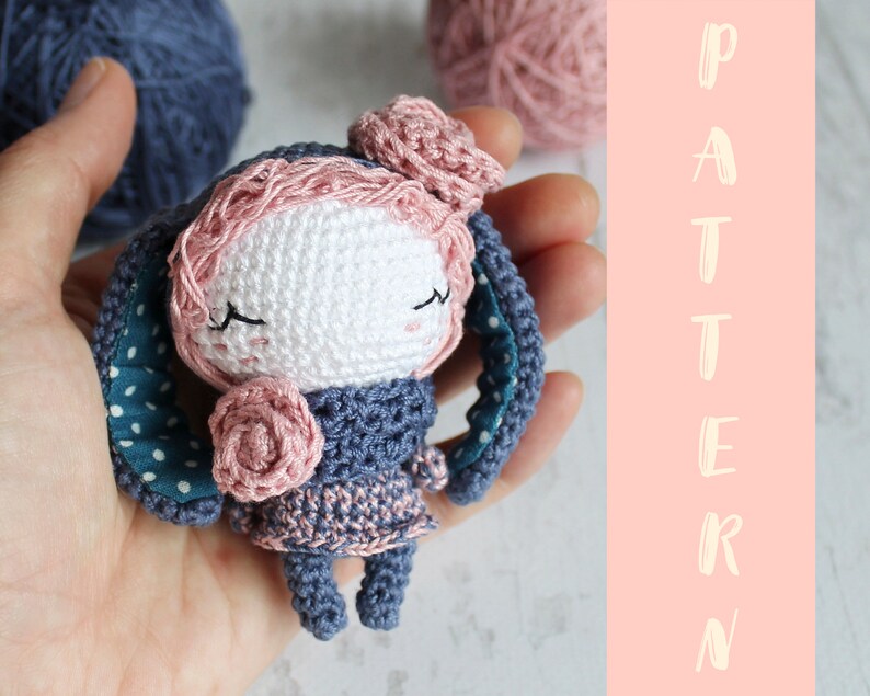 K&ouml;nnte beinhalten: Eine geh&auml;kelte Puppe aus rosa und blauem Garn. Die Puppe hat ein wei&szlig;es Gesicht mit geschlossenen Augen und eine rosa Blume auf der Brust. Die Puppe h&auml;lt eine blau-wei&szlig; gepunktete Decke.