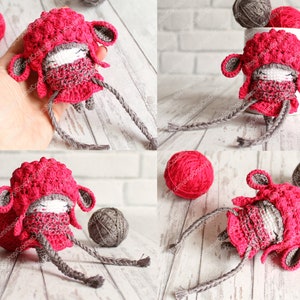 May include: A crocheted amigurumi doll wearing a pink hat and scarf. The doll has gray yarn for arms and legs. The doll is sitting on a white wooden surface.