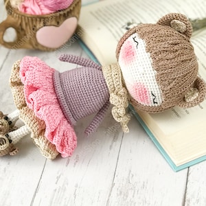 May include: A crocheted doll with brown bear ears, wearing a purple dress and a pink tutu, lying on a book. The doll has a white face with pink cheeks and closed eyes.