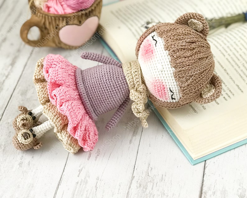 May include: A crocheted doll with brown bear ears, wearing a purple dress and a pink tutu, lying on a book. The doll has a white face with pink cheeks and closed eyes.