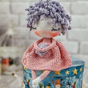 May include: A crocheted doll with a pink dress and purple hair sitting on a blue and white gift box with stars.