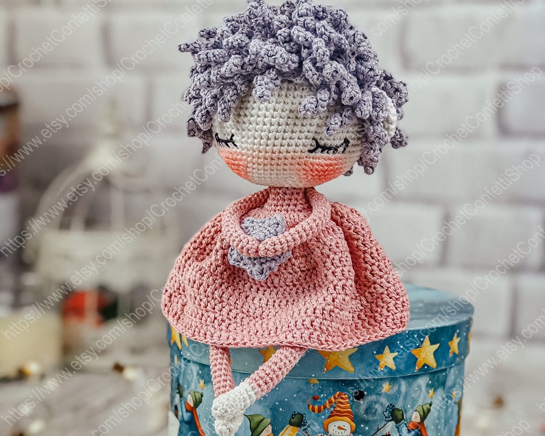 May include: A crocheted doll with a pink dress and purple hair sitting on a blue and white gift box with stars.