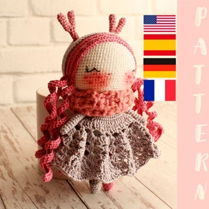 May include: Crocheted amigurumi doll with pink yarn hair, a pink scarf, and a gray dress. The doll has antlers and closed eyes. The doll is sitting on a wooden surface.