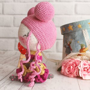 Pu&ograve; includere: Una bambola all'uncinetto con i capelli rosa, un cappello rosa e un vestito colorato. La bambola &egrave; seduta su una superficie bianca.