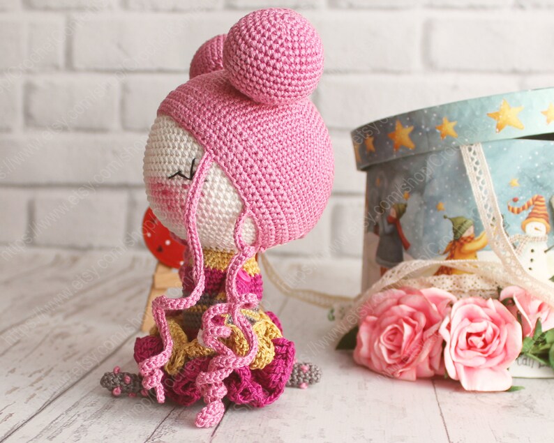 Pu&ograve; includere: Una bambola all'uncinetto con i capelli rosa, un cappello rosa e un vestito colorato. La bambola &egrave; seduta su una superficie bianca.