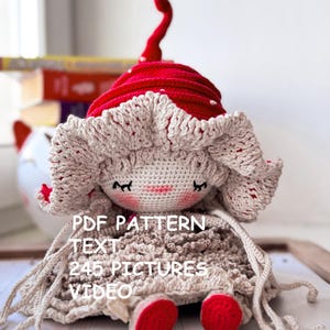Op de afbeelding: Een handgemaakte gehaakte pop met een rode hoed en bijpassende schoenen. De pop heeft een beige gezicht en jurk, met een witte, kantachtige rand rond de hoed en jurk. Het gezicht van de pop heeft gesloten ogen, roze wangen en een lachende mond. Tekst op de pop: PDF PATTERN TEXT 245 PICTURES VIDEO.
