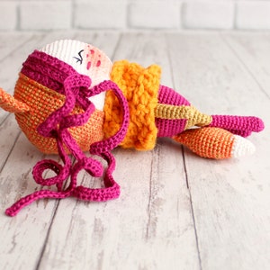 May include: A handmade crochet fox doll in shades of orange, pink, and white. The doll has a round head with closed eyes, a long tail, and a scarf. The fox is lying down on a light-colored wooden surface.
