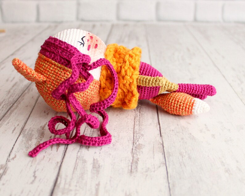May include: A handmade crochet fox doll in shades of orange, pink, and white. The doll has a round head with closed eyes, a long tail, and a scarf. The fox is lying down on a light-colored wooden surface.