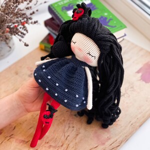 K&ouml;nnte beinhalten: H&auml;kelpuppe mit langen schwarzen Haaren, einem blauen Kleid mit wei&szlig;en Punkten und roten Schuhen. Die Puppe hat einen Marienk&auml;fer auf dem Kopf und h&auml;lt einen Marienk&auml;fer in der Hand.