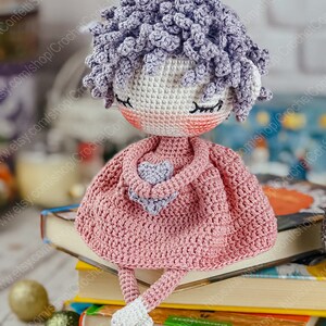 May include: A crocheted doll with purple hair, wearing a pink dress and holding a purple heart. The doll is sitting on a stack of books.