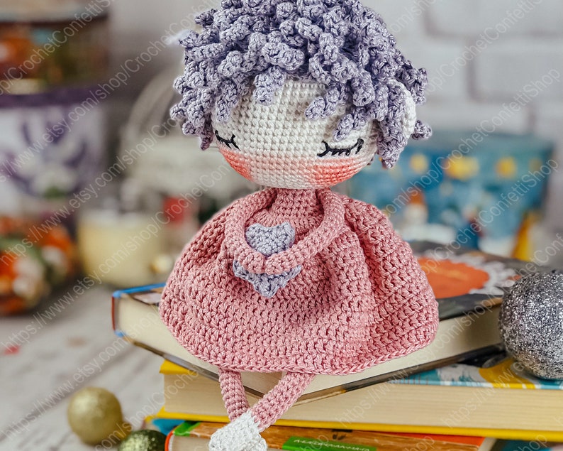 May include: A crocheted doll with purple hair, wearing a pink dress and holding a purple heart. The doll is sitting on a stack of books.