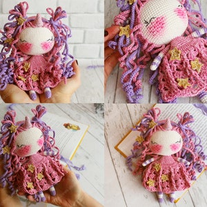 Puede incluir: Una mu&ntilde;eca de unicornio de crochet hecha con hilo rosa y morado, que lleva un vestido rosa con estrellas doradas. La mu&ntilde;eca tiene una cara blanca con mejillas rosadas y los ojos cerrados.