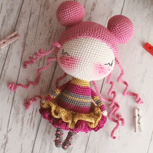 Pu&ograve; includere: Una bambola all'uncinetto con i capelli rosa, un vestito a righe giallo e rosa e una gonna a balze gialla. La bambola ha gli occhi chiusi e un blush rosa sulle guance.