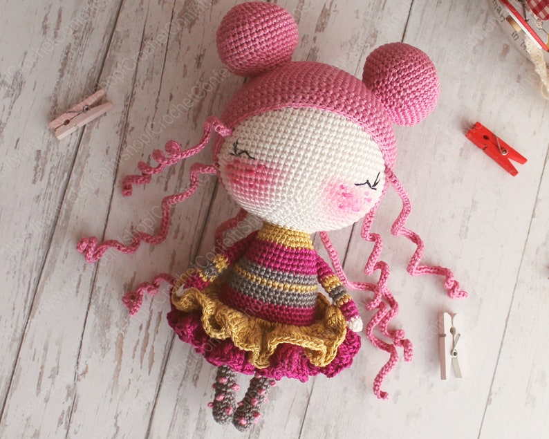 Pu&ograve; includere: Una bambola all'uncinetto con i capelli rosa, un vestito a righe giallo e rosa e una gonna a balze gialla. La bambola ha gli occhi chiusi e un blush rosa sulle guance.