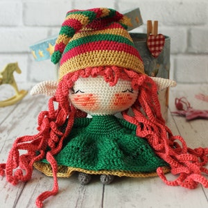 Peut inclure: Une poup&eacute;e elfe en crochet avec de longs cheveux roux, portant une robe verte et un chapeau ray&eacute;. La poup&eacute;e est assise sur une surface en bois blanc.