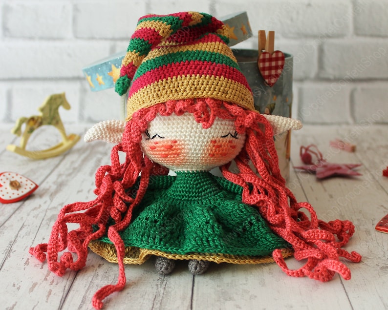 Peut inclure: Une poup&eacute;e elfe en crochet avec de longs cheveux roux, portant une robe verte et un chapeau ray&eacute;. La poup&eacute;e est assise sur une surface en bois blanc.