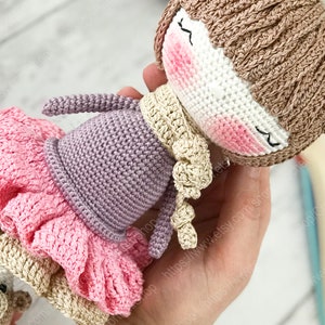 May include: A crocheted doll wearing a pink and brown dress, a purple sweater, and a brown hat with bear ears. The doll has a white face with pink cheeks and closed eyes.
