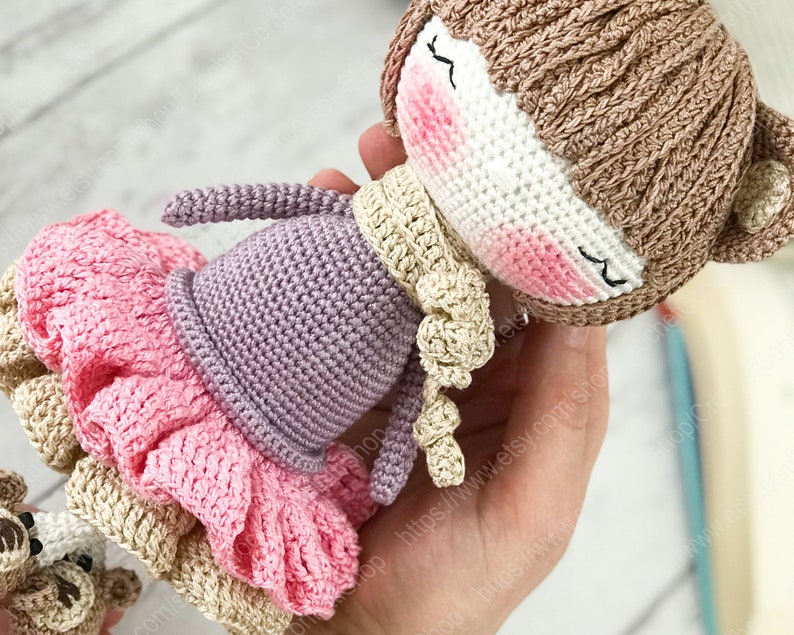 May include: A crocheted doll wearing a pink and brown dress, a purple sweater, and a brown hat with bear ears. The doll has a white face with pink cheeks and closed eyes.