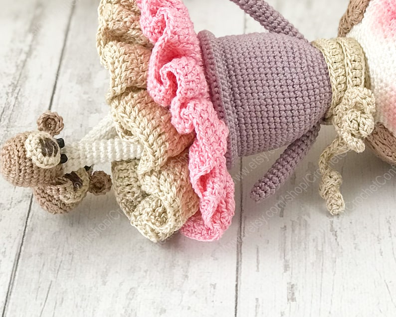 May include: A crocheted doll wearing a pink and beige dress with a purple top and a brown crocheted monkey attached to its arm.