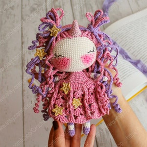 Puede incluir: Una mu&ntilde;eca de unicornio de crochet con pelo de lana rosa y morado, un vestido rosa con estrellas doradas y un cuerno rosa. La mu&ntilde;eca est&aacute; siendo sostenida por una mano.
