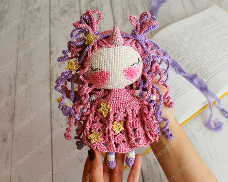 Puede incluir: Una mu&ntilde;eca de unicornio de crochet con pelo de lana rosa y morado, un vestido rosa con estrellas doradas y un cuerno rosa. La mu&ntilde;eca est&aacute; siendo sostenida por una mano.