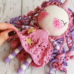Puede incluir: Una mu&ntilde;eca de unicornio de crochet con un vestido rosa y pelo morado. El vestido tiene detalles de estrellas doradas.