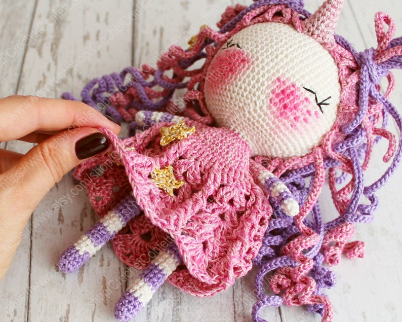 Puede incluir: Una mu&ntilde;eca de unicornio de crochet con un vestido rosa y pelo morado. El vestido tiene detalles de estrellas doradas.