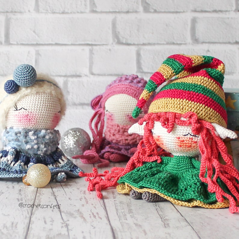 Peut inclure: Trois poup&eacute;es en crochet avec des tenues et des cheveux de diff&eacute;rentes couleurs. La poup&eacute;e &agrave; gauche porte une tenue bleu et blanc avec un chapeau blanc. La poup&eacute;e du milieu porte une tenue rose et blanc avec un chapeau rose. La poup&eacute;e &agrave; droite porte une tenue verte et dor&eacute;e avec un chapeau rouge et vert. Les poup&eacute;es sont assises sur une surface en bois blanc. @crochetconfetti