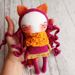 May include: A handmade crochet doll with a fox-like design. The doll features a white face, pink cheeks, and closed eyes. It wears a pink dress, an orange scarf, and has curly pink hair. The doll's ears are orange, and the overall design is whimsical.