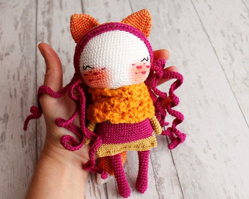 May include: A handmade crochet doll with a fox-like design. The doll features a white face, pink cheeks, and closed eyes. It wears a pink dress, an orange scarf, and has curly pink hair. The doll's ears are orange, and the overall design is whimsical.