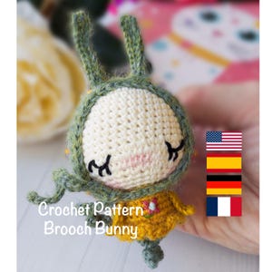 May include: A handmade crochet bunny brooch with a cream-colored head, green ears, and a yellow dress. The brooch has embroidered facial features and the text "Crochet Pattern Brooch Bunny".