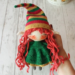 Peut inclure: Une poup&eacute;e elfe en crochet avec des cheveux en laine rouge, une robe verte et un chapeau ray&eacute; rouge, jaune et vert.