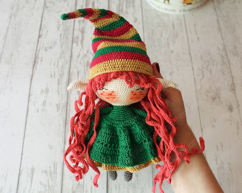 Peut inclure: Une poup&eacute;e elfe en crochet avec des cheveux en laine rouge, une robe verte et un chapeau ray&eacute; rouge, jaune et vert.