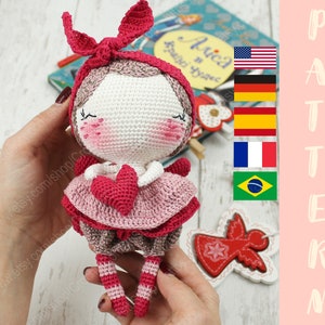 May include: Crocheted doll with pink and brown clothing, holding a pink heart. The doll has a pink bow and brown hair. The doll is being held by a hand.