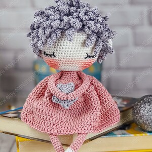 May include: A crocheted doll with gray hair, pink dress, and a gray star in her hands. The doll is sitting on a stack of books.