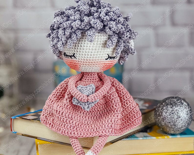 May include: A crocheted doll with gray hair, pink dress, and a gray star in her hands. The doll is sitting on a stack of books.