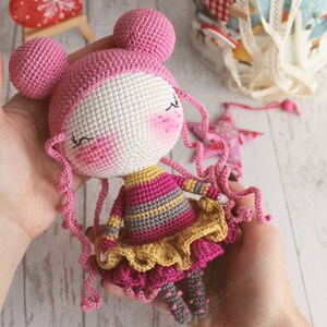 Pu&ograve; includere: Una bambola all'uncinetto con capelli rosa, un cappello rosa e bianco, un vestito a righe rosa e grigio e una gonna a balze gialla.